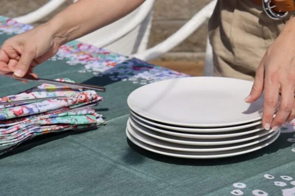 Hands setting a stack of white plates on the dark green tablecloth beside folded floral napkins.